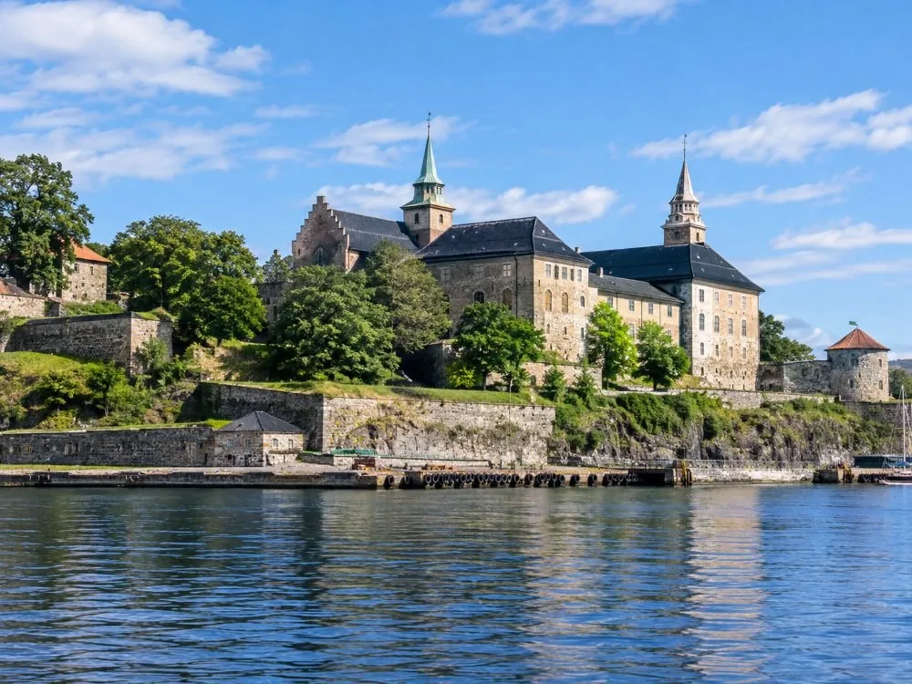 Die Festung Akershus vom Fjord aus gesehen: helle Steinmauern und steile Dächer, die auf einer grünen Landzunge direkt über dem Hafenwasser stehen, mit der Kaipromenade unter den Wallanlagen.