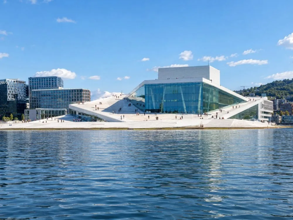 Das Opernhaus von Oslo auf der anderen Seite von Bjørvika: ein flaches weißes Gebäude aus hellem Stein und Glas, das sich direkt ins Wasser neigt, mit kleinen Menschen auf seinem schrägen Dach.