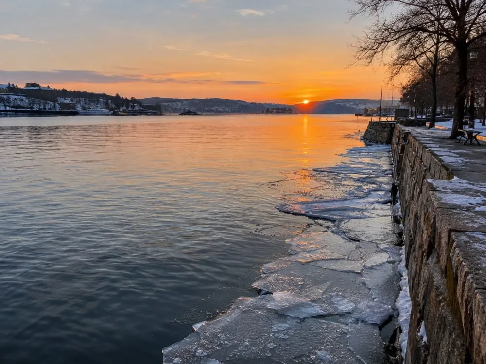 Der innere Oslofjord an einem WinterNachmittag: flaches graues Wasser, dünnes Eis am Kai und das tiefe orangefarbene Licht einer Sonne, die bereits am frühen Nachmittag untergeht.